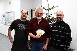 Drei Männer stehen in einem Gebäude vor einem Weihnachtsbaum. Der Herr in der Mitte hält einen Fächer aus Geldscheinen in der Hand.