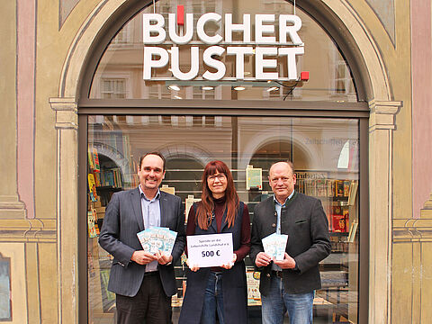 Johannes Fauth, Kerstin Glöckner und Hans-Peter Summer stehen vor dem Eingang der Buchhandlung und halten Weihnachtskarten und ein Schild mit der Aufschrift: Spende an die Lebenshilfe Landshut 500 Euro